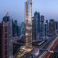 A tall, modern skyscraper in an urban skyline at dusk, surrounded by other high-rise buildings and busy highways with streaking car lights.