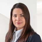 Portrét Zuzany Vaškovičové. Žena s rovnými hnědými vlasy, v bílé košili a tmavém saku, stojí před světlým pozadím s klidným výrazem ve tváři.