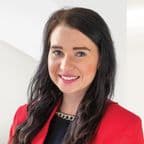 Portrait of Gabriela Szőke. A young woman with long dark hair, wearing a black top with a chain detail and a red blazer. She is smiling and standing in front of a bright background.