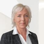 Usměvavá HR specialistka v saku stojí před světlou stěnou a vyzařuje přátelský a vstřícný dojem.