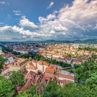 Luftaufnahme der historischen Altstadt von Graz, Österreich, aufgenommen vom Schlossberg. Im Vordergrund sind üppige grüne Vegetation und pink blühende Blumen zu sehen. Die Stadt erstreckt sich mit ihren charakteristischen roten Ziegeldächern und historischen Gebäuden bis zum Horizont. Markante Kirchtürme und historische Architektur prägen das Stadtbild. Im Hintergrund erheben sich die grünen Hügel der steirischen Berglandschaft unter einem dramatischen Himmel mit weißen Wolkenformationen. Die Aufnahme zeigt den Kontrast zwischen der dicht bebauten Altstadt und der modernen Stadtentwicklung am Horizont.
