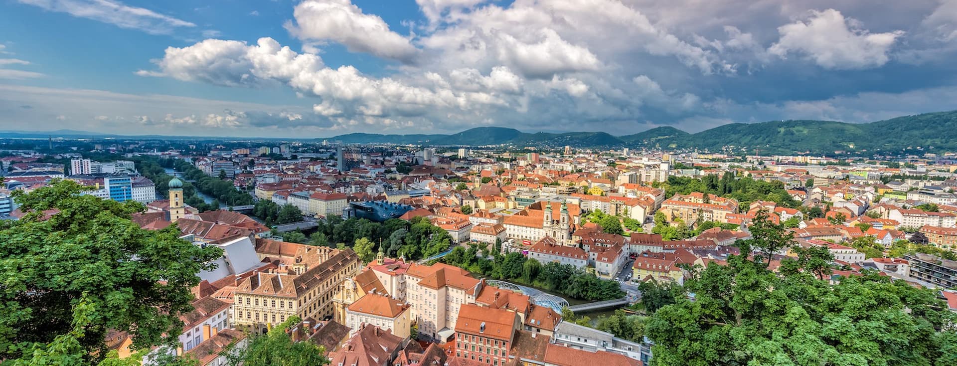 Luftaufnahme der historischen Altstadt von Graz, Österreich, aufgenommen vom Schlossberg. Im Vordergrund sind üppige grüne Vegetation und pink blühende Blumen zu sehen. Die Stadt erstreckt sich mit ihren charakteristischen roten Ziegeldächern und historischen Gebäuden bis zum Horizont. Markante Kirchtürme und historische Architektur prägen das Stadtbild. Im Hintergrund erheben sich die grünen Hügel der steirischen Berglandschaft unter einem dramatischen Himmel mit weißen Wolkenformationen. Die Aufnahme zeigt den Kontrast zwischen der dicht bebauten Altstadt und der modernen Stadtentwicklung am Horizont.