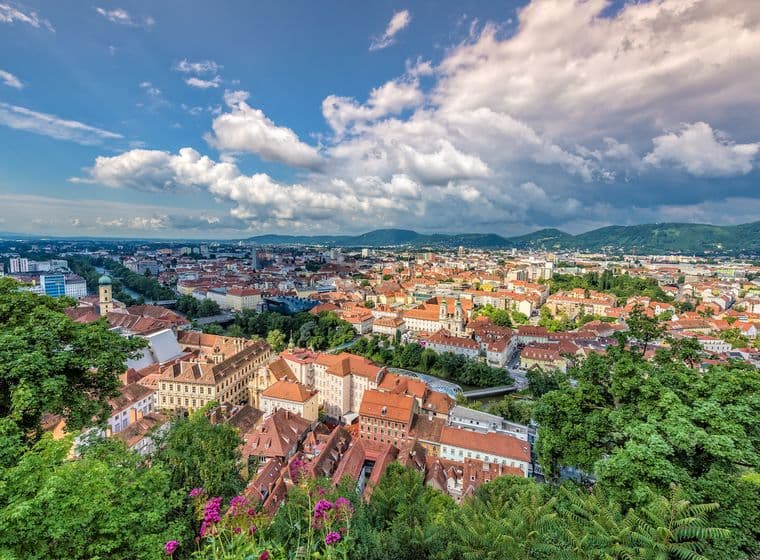 Luftaufnahme der historischen Altstadt von Graz, Österreich, aufgenommen vom Schlossberg. Im Vordergrund sind üppige grüne Vegetation und pink blühende Blumen zu sehen. Die Stadt erstreckt sich mit ihren charakteristischen roten Ziegeldächern und historischen Gebäuden bis zum Horizont. Markante Kirchtürme und historische Architektur prägen das Stadtbild. Im Hintergrund erheben sich die grünen Hügel der steirischen Berglandschaft unter einem dramatischen Himmel mit weißen Wolkenformationen. Die Aufnahme zeigt den Kontrast zwischen der dicht bebauten Altstadt und der modernen Stadtentwicklung am Horizont.