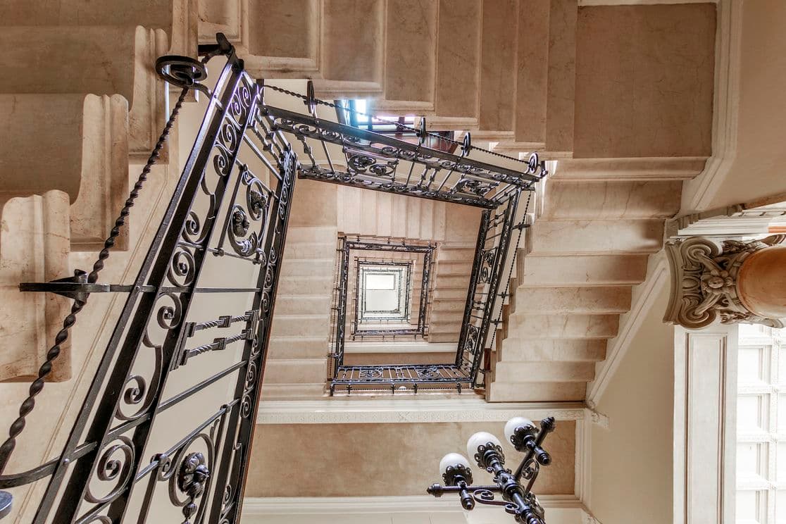 Blick nach oben auf eine Wendeltreppe mit verziertem schwarzen Geländer und beigen Steinstufen, die ein geometrisches Muster bilden.