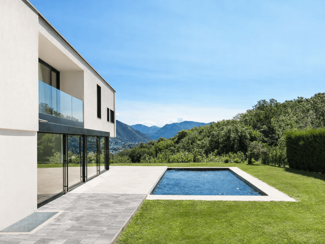 Modern house with large windows, patio, and a rectangular pool. Surrounded by lush greenery and a backdrop of distant mountains under a clear blue sky.