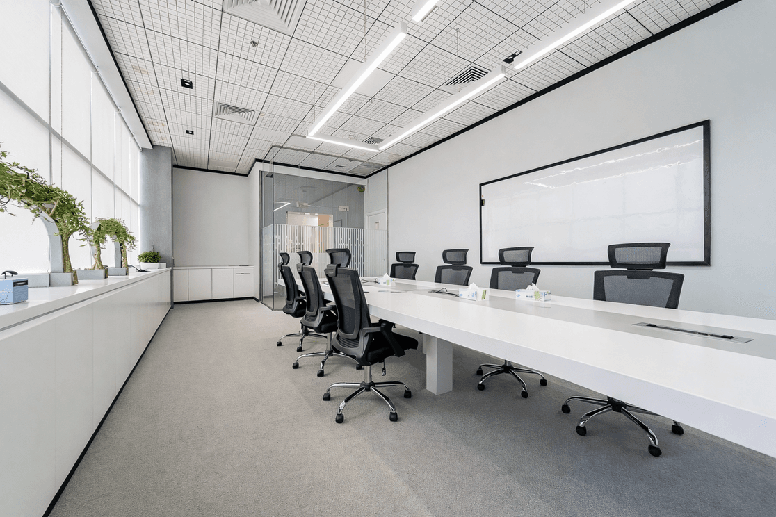 Minimalist conference room with long white table, eight black chairs, a large whiteboard, and potted plants on window ledge.