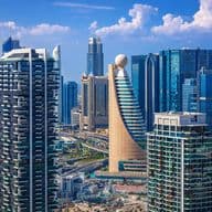 Aerial view of a modern city skyline with tall, diverse skyscrapers, highways, and a clear blue sky with scattered clouds.