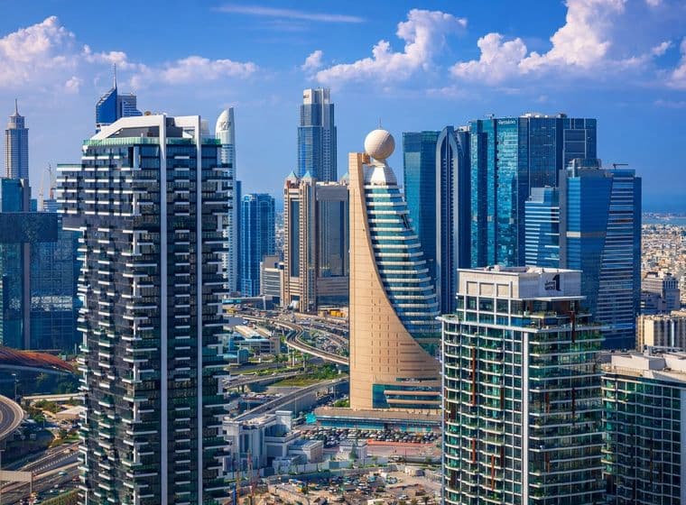 Luftaufnahme einer modernen Skyline mit hohen, vielfältigen Wolkenkratzern, Schnellstraßen und einem klaren blauen Himmel mit vereinzelten Wolken.