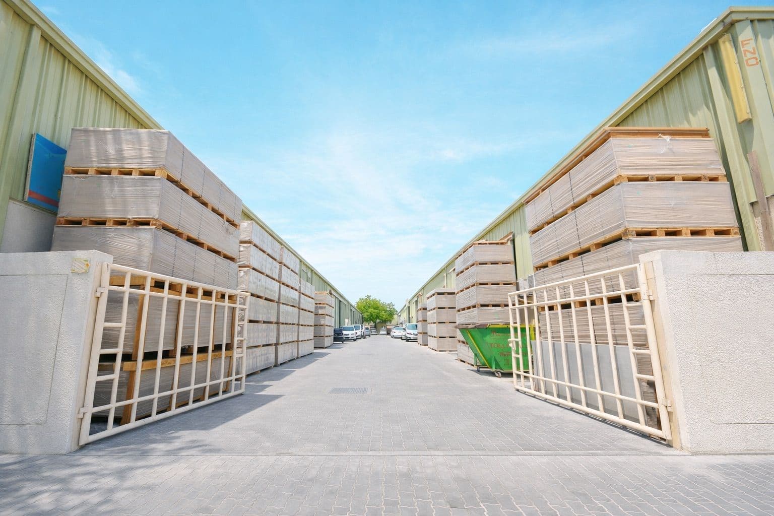 Paved passageway between two large industrial warehouses storing stacks of wooden pallets beneath a clear blue sky.