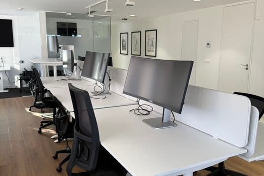 Modern office with white desks, black chairs, and large monitors. Hardwood floor and framed art on the walls. Bright and minimalist design.