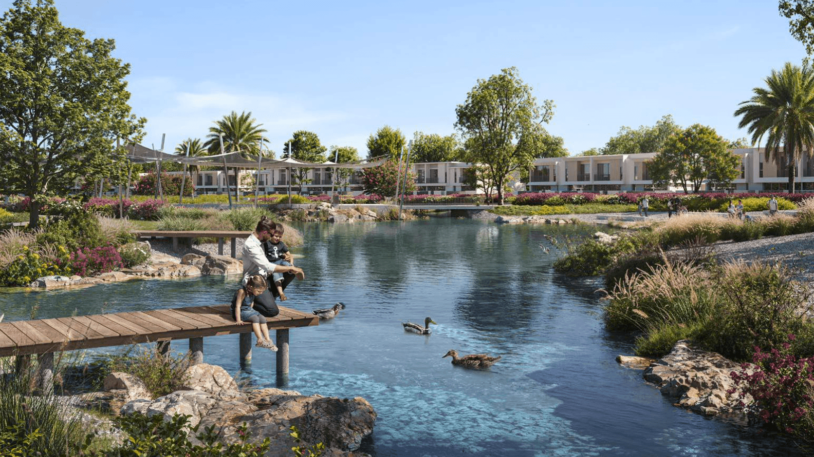 Personen sitzen auf einem Holzsteg an einem Teich mit Enten, umgeben von Gruenflaechen und modernen Hausern im Hintergrund.