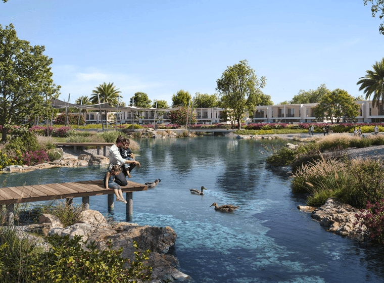 People sitting on a wooden dock by a pond with ducks, surrounded by greenery and modern houses in the background.