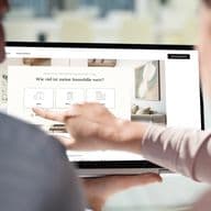 Zwei Personen nutzen ein Tablet, um eine Immobilien-Website zu betrachten, mit einer Frage auf Deutsch zum Immobilienwert. Eine Person zeigt auf den Bildschirm.