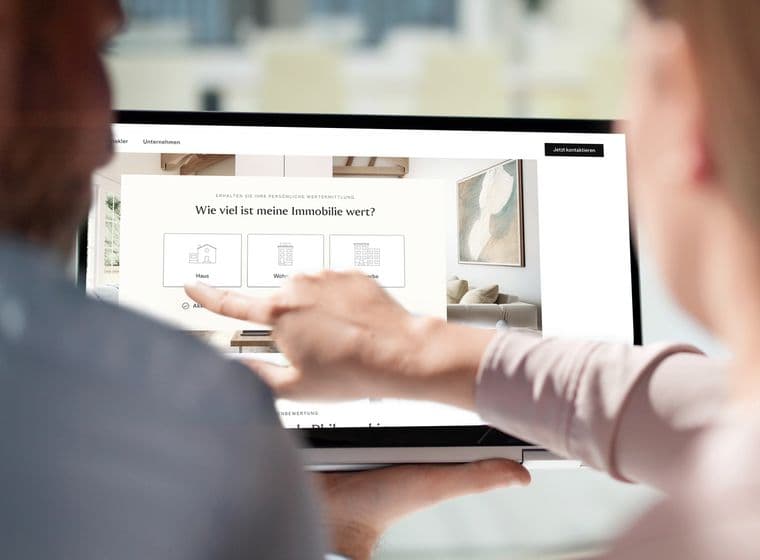 Zwei Personen nutzen ein Tablet, um eine Immobilien-Website zu betrachten, mit einer Frage auf Deutsch zum Immobilienwert. Eine Person zeigt auf den Bildschirm.