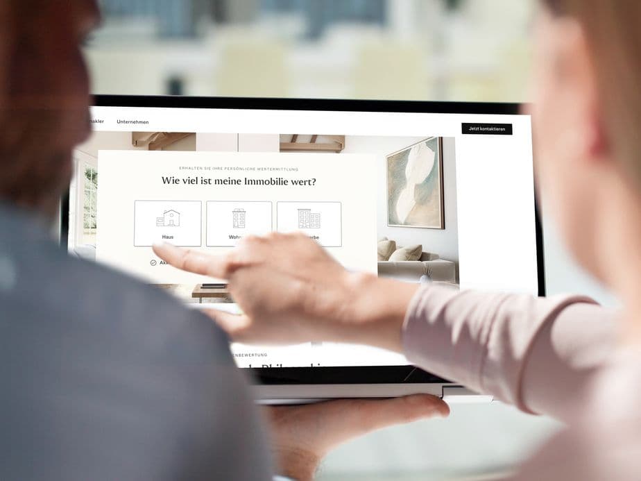 Two people using a tablet to view a real estate website with a question in German about property value. One person points at the screen.