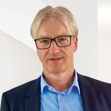 Headshot of a man with gray hair and glasses, wearing a blue shirt and navy blazer.