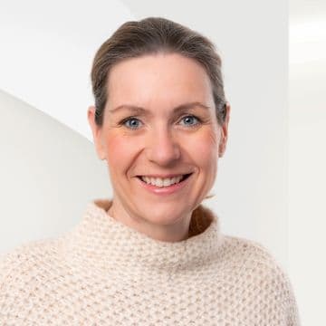 Woman with light brown hair in a bun, smiling, wearing a textured beige sweater against a light background.