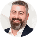 Headshot of a smiling man with a beard, wearing a navy suit and white shirt, set against a white background.