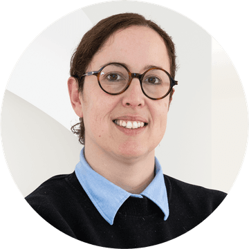 A woman with glasses, wearing a blue collared shirt and black sweater, smiles against a neutral background.
