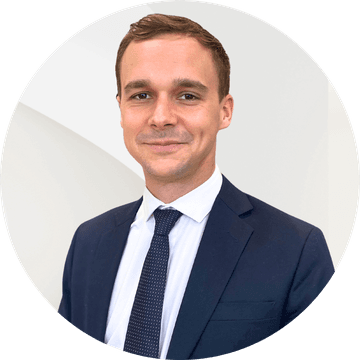 Smiling man wearing a dark suit jacket, white shirt, and dark tie, in front of a plain white background.