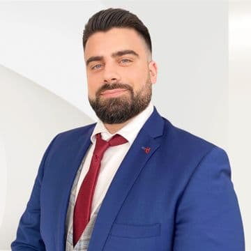 Man in a blue suit and red tie, with a neatly groomed beard and styled hair, stands confidently against a light, abstract background.