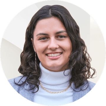 Close-up of a smiling young woman with dark curly hair wearing a light turtleneck, gray blazer, gold chain necklace and a dangling earring.