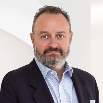 Headshot of a man with a beard, wearing a blue striped shirt and a dark blazer, against a white background.