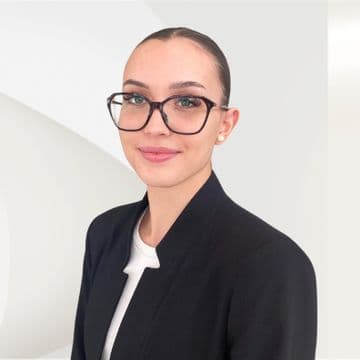 A woman with glasses and a neat bun, wearing a black blazer over a white top, smiles against a light, abstract background.