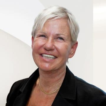 Smiling woman with short blonde hair, wearing a black jacket and gold necklace, against a light background.