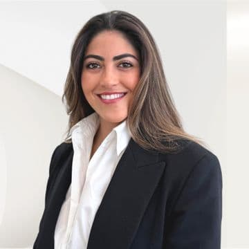 Smiling woman with long hair wearing a black blazer and white shirt, standing against a light background.