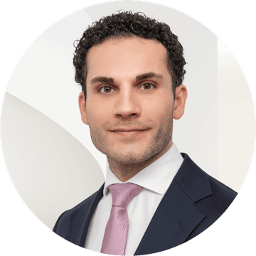 Man with curly hair in a suit and tie, posing against a neutral background, looking directly at the camera with a slight smile.
