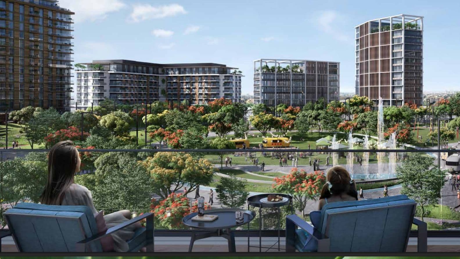 Two people seated on a balcony overlooking a vibrant urban park with lush greenery, fountains, and modern buildings under a clear sky.