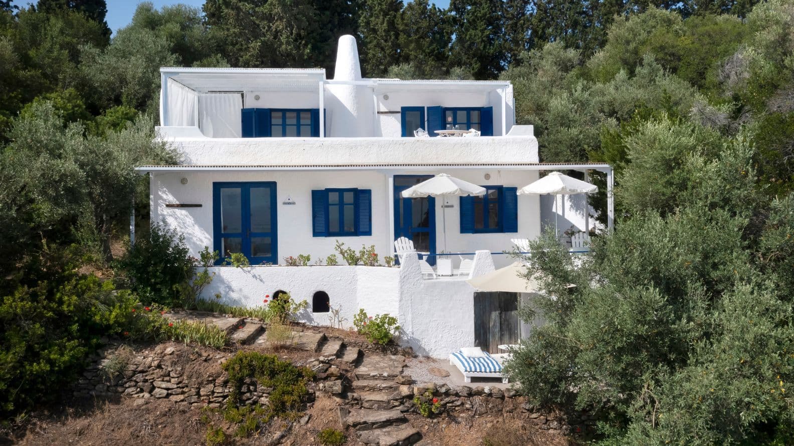 Two-story white villa with traditional blue shutters and terraces, surrounded by mature trees.