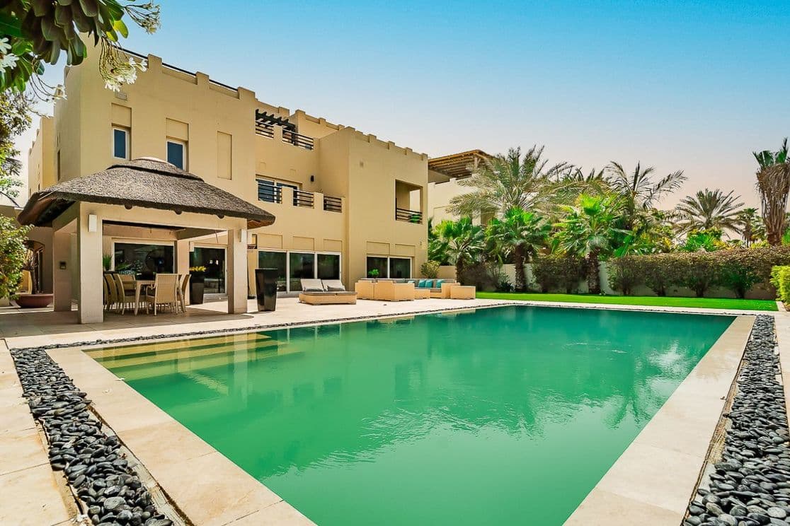 Luxuriöse Villa mit überdachtem Gazebo, Pool mit Natursteinumrandung, üppigem Grün und Palmen unter blauem Himmel.