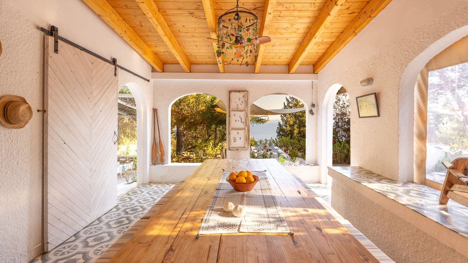 Semi-outdoor dining space featuring an exposed beam ceiling, patterned floor tiles, a long wooden table, and arched openings with sea views.