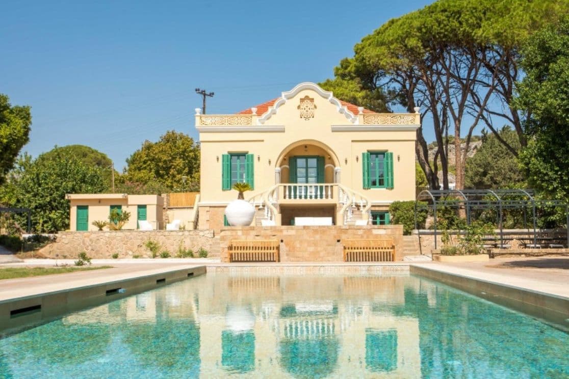 Elegant neoclassical villa with a private swimming pool, surrounded by lush greenery and tall pine trees under a clear blue sky.