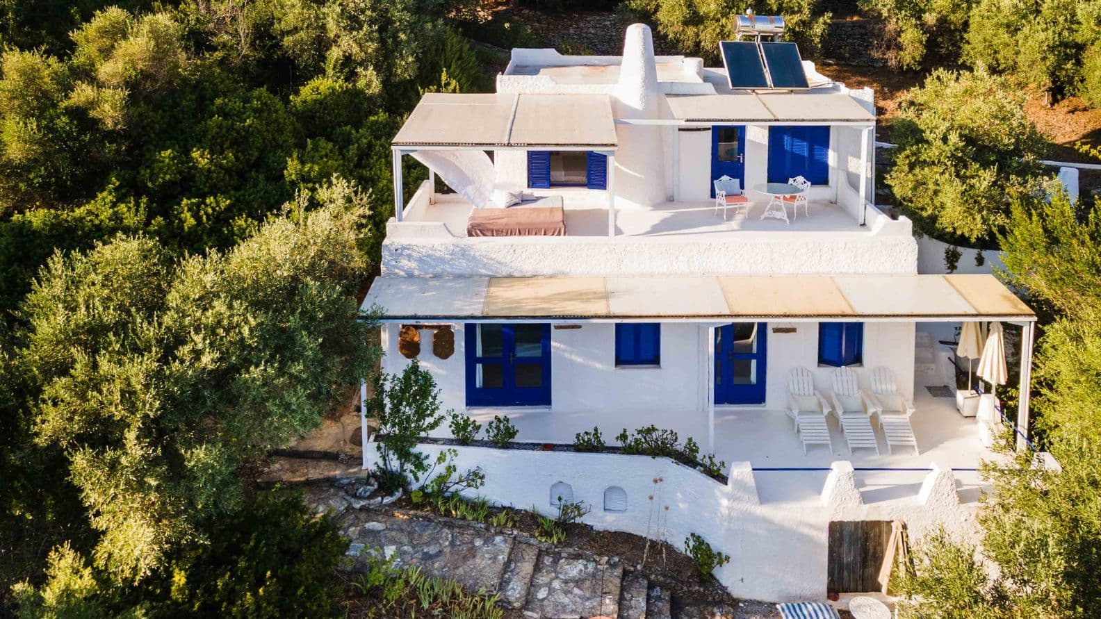 Two-story white villa with traditional blue shutters, terraces, and solar panels, surrounded by mature trees.