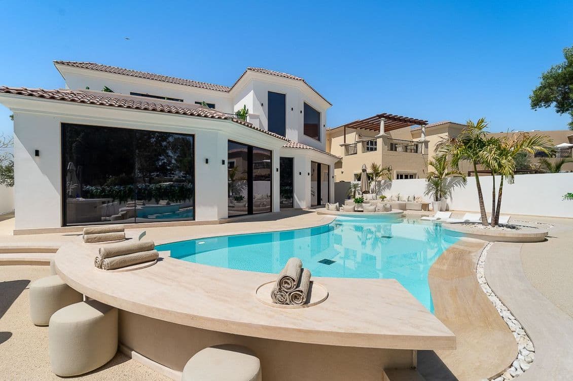 Luxurious modern villa with a curved pool, sun loungers, and palm trees, set against a clear blue sky.