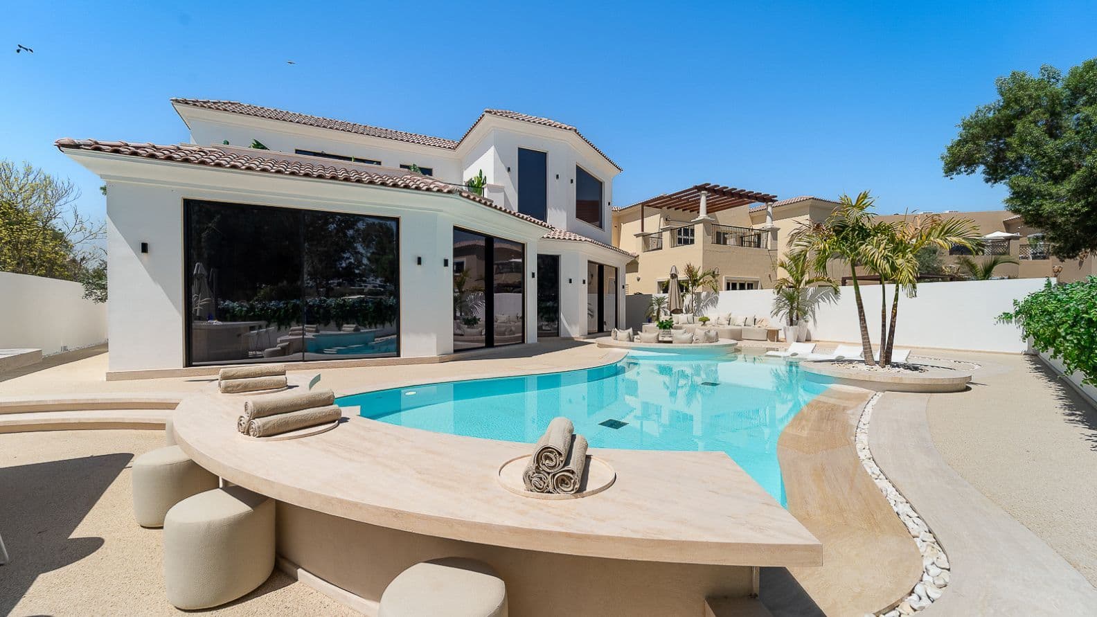 Luxurious modern villa with a curved pool, sun loungers, and palm trees, set against a clear blue sky.