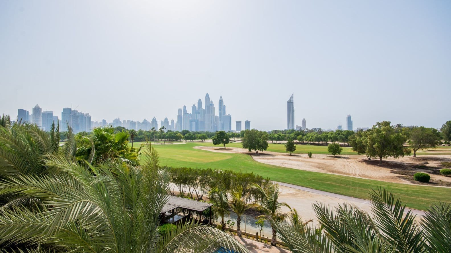 Aussicht auf die Skyline der Dubai Marina mit modernen Wolkenkratzern, einem üppig grünen Golfplatz und Palmen im Vordergrund unter blauem Himmel.