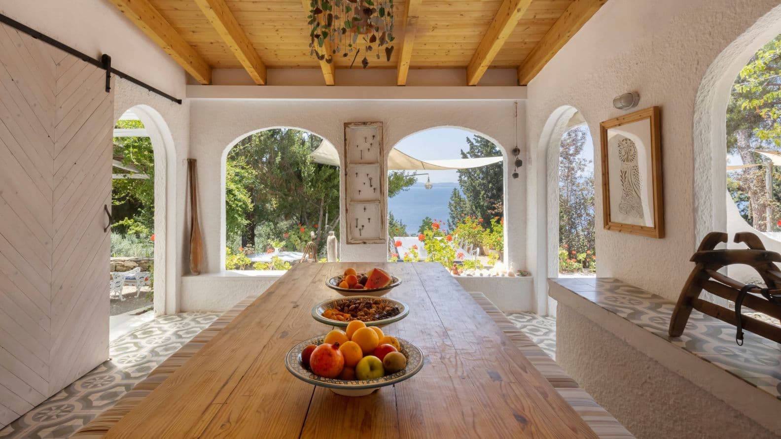 Semi-outdoor dining space featuring an exposed beam ceiling, patterned floor tiles, a long wooden table, and arched openings with sea views.