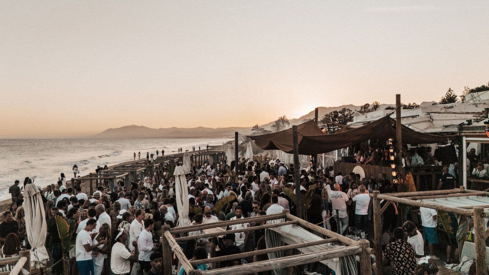 Fiesta en la playa en Marbella con mucha gente junto al mar en un chiringuito al atardecer, con las montañas de fondo.