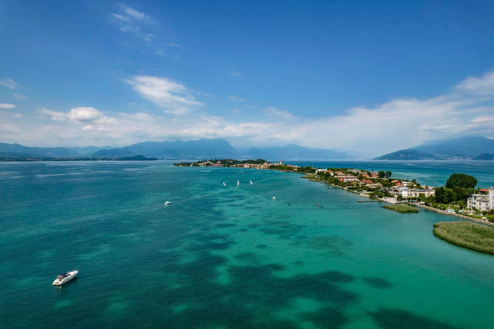 Veduta aerea della penisola di Sirmione che si estende nelle acque turchesi del Lago di Garda, con barche sparse e le montagne sullo sfondo in una giornata serena.