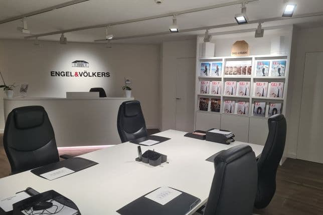 Modern office interior with a reception desk, branded "Engel & Völkers," chairs, a conference table with folders, and magazine displays on shelves.