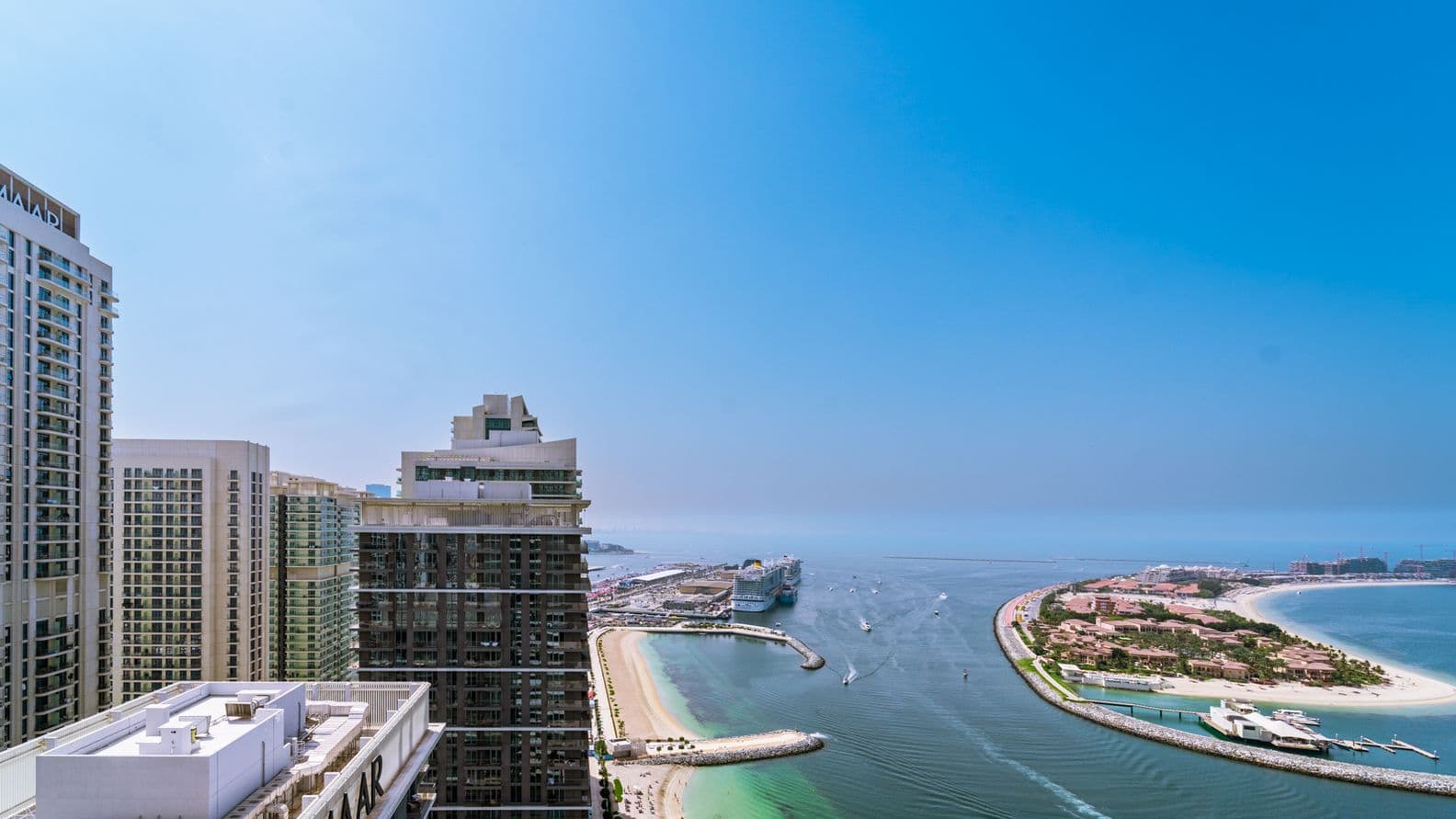 Aerial view of a coastal cityscape with high-rise buildings, a marina, and a curved sandy beach under a clear blue sky.