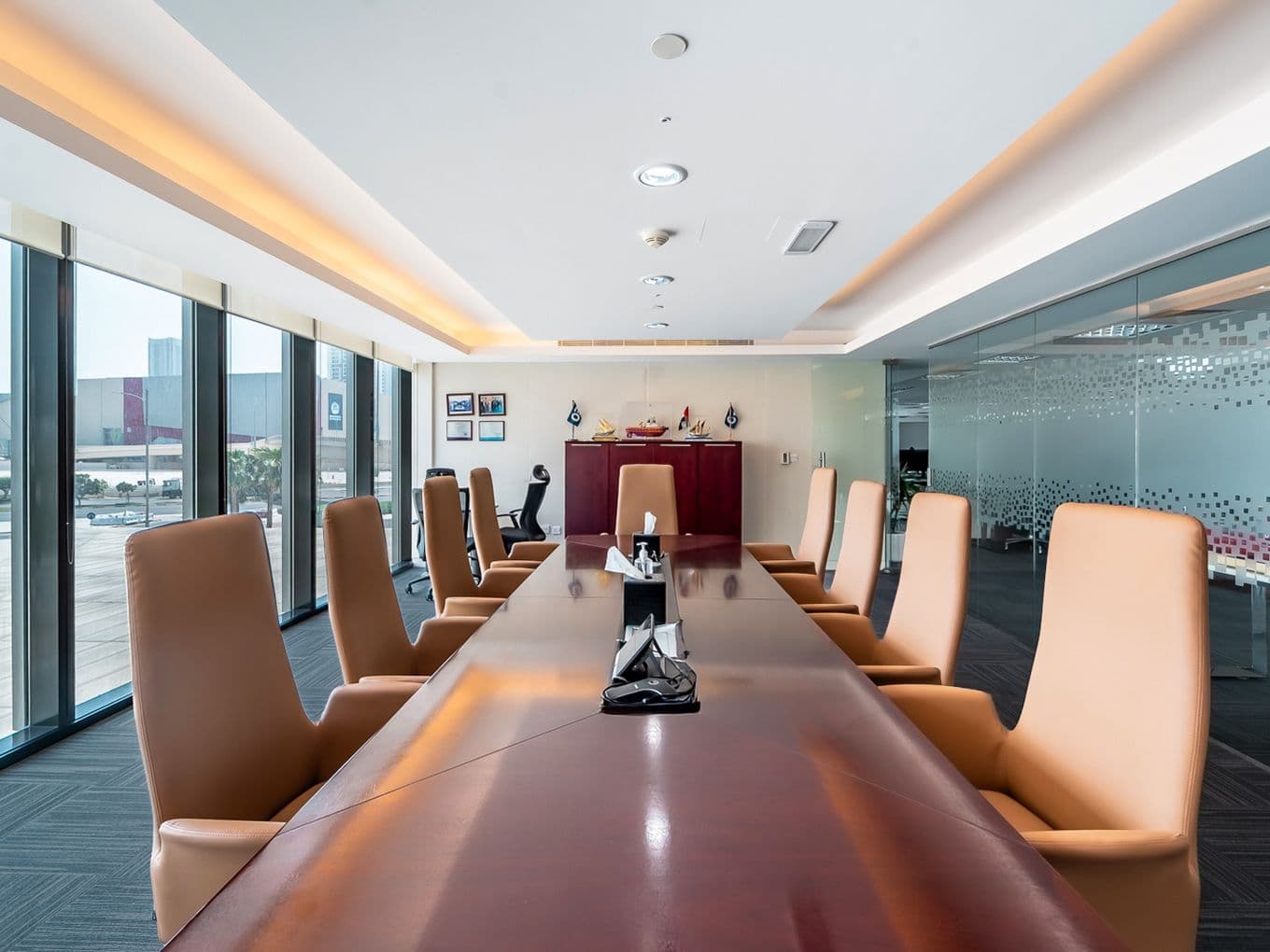 Heller moderner Sitzungssaal mit einem langen glänzenden Konferenztisch aus Holz, beigefarbenen Stühlen mit hoher Rückenlehne, raumhohen Fenstern und einer Glaswand.