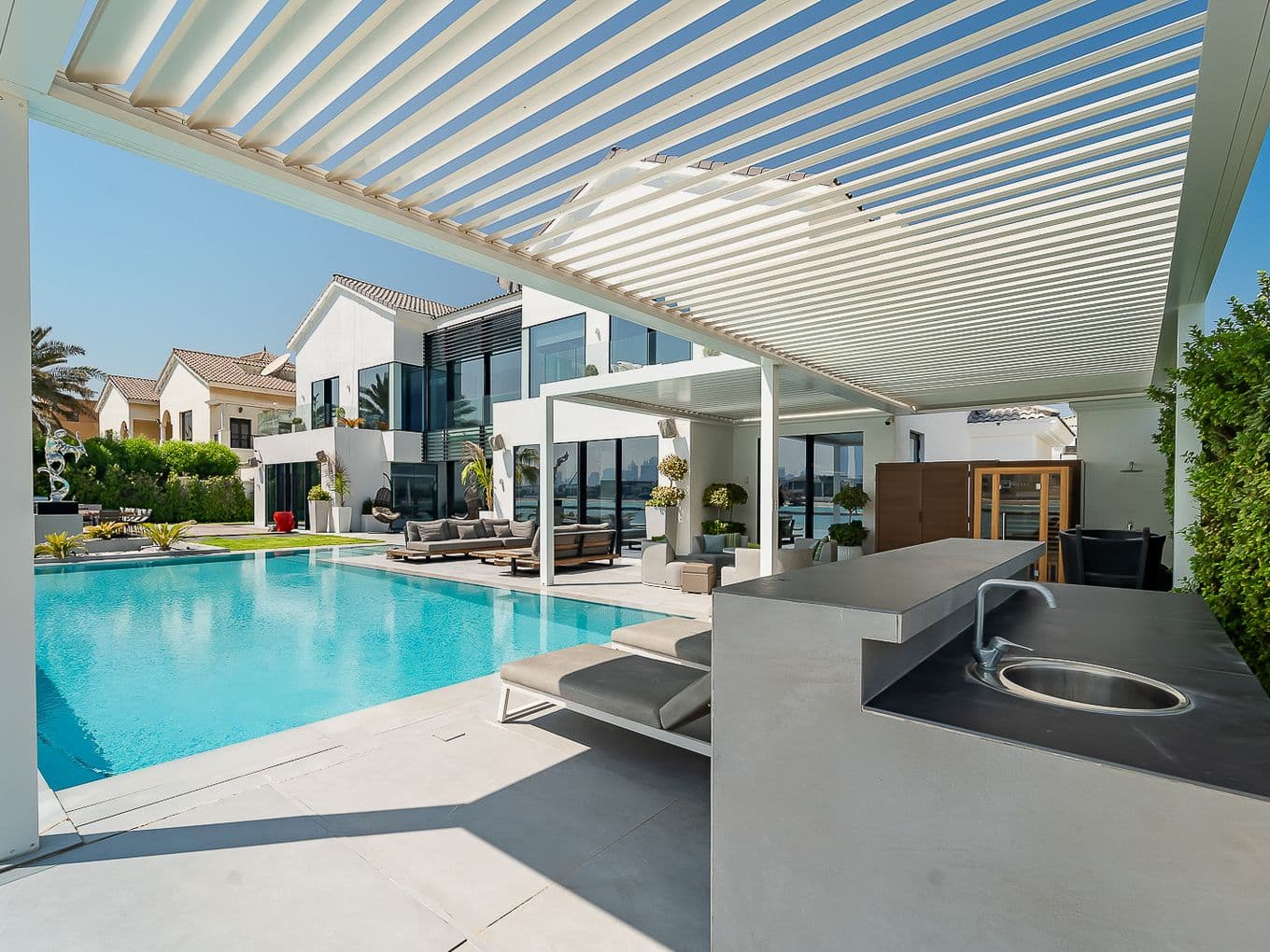 Moderner Hinterhof mit Pool, Pergola, Sonnenliegen und Außenküche. Im Hintergrund ein zeitgenössisches Haus und ein gepflegter Garten.