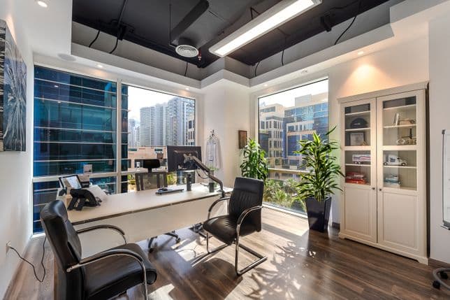 Bright modern corner office with white desk, two black chairs, tall plants and city-view windows.