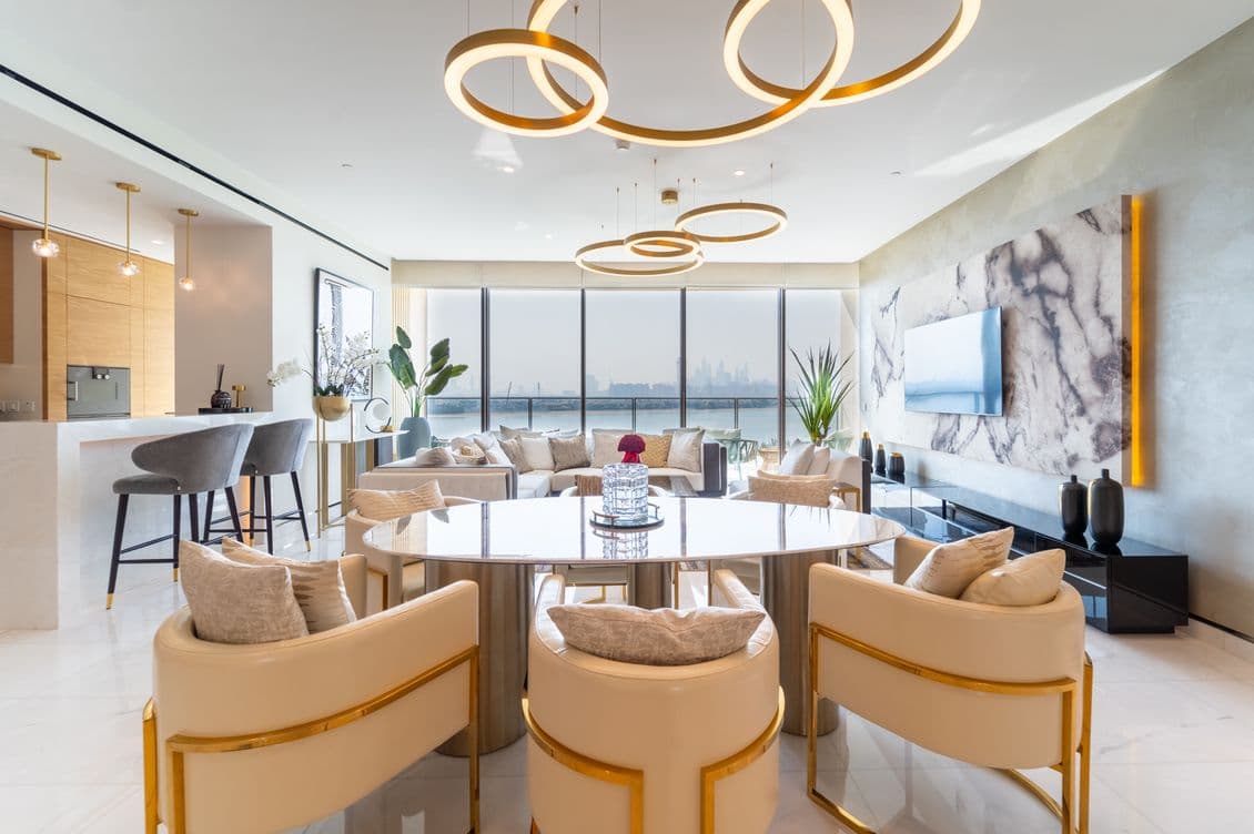 Modern living room with circular chandelier, marble wall, large windows, round dining table, cream chairs, and a view of the city skyline.