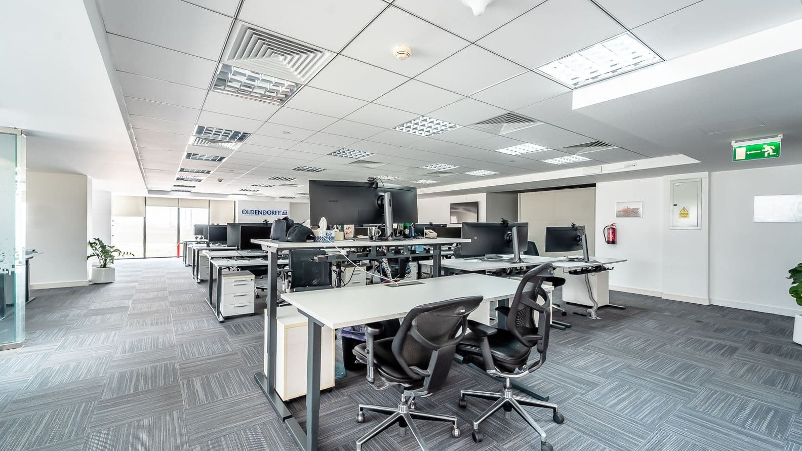 Helles Großraumbüro mit Reihen von weißen Schreibtischen, schwarzen ergonomischen Stühlen und Monitoren auf grauem Teppich unter einer abgehängten Decke.